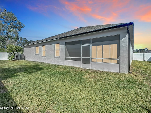 back of house at dusk featuring a sunroom, roof with shingles, fence, a yard, and stucco siding