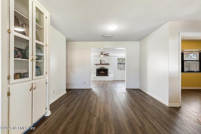 unfurnished living room featuring baseboards, ceiling fan, dark wood-type flooring, a textured ceiling, and a brick fireplace