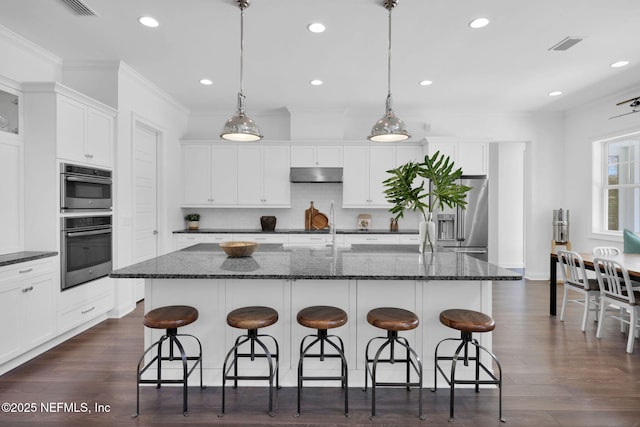 kitchen with under cabinet range hood, ornamental molding, white cabinets, stainless steel appliances, and a kitchen island with sink