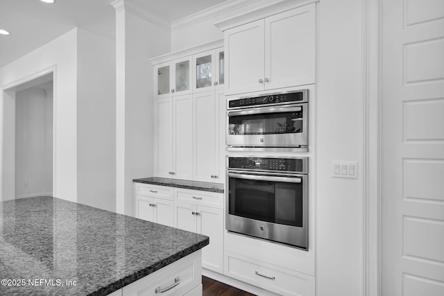 kitchen with stainless steel double oven, dark stone countertops, white cabinetry, and ornamental molding