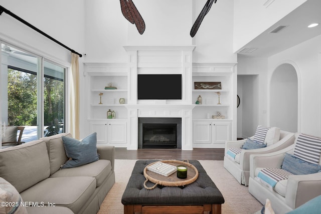 living area with visible vents, built in shelves, a towering ceiling, wood finished floors, and a glass covered fireplace