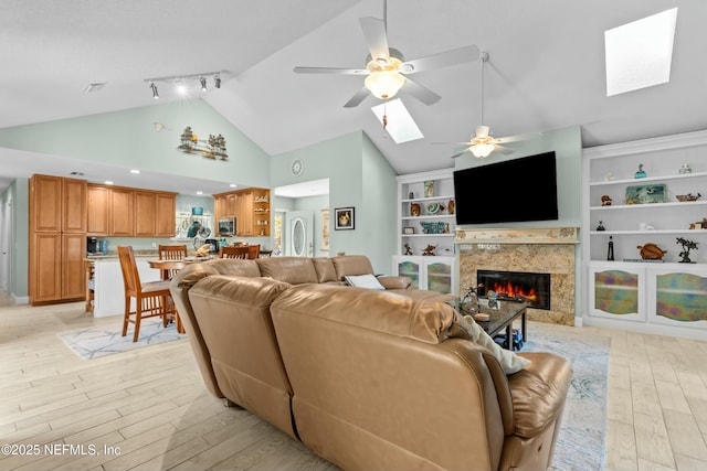 living room with high vaulted ceiling, a skylight, a fireplace, a ceiling fan, and light wood finished floors