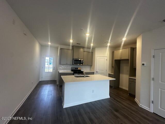 kitchen featuring dark wood-style floors, a center island with sink, gray cabinetry, appliances with stainless steel finishes, and backsplash