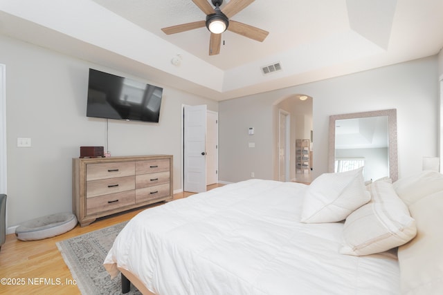 bedroom with light wood-type flooring, a tray ceiling, arched walkways, and visible vents