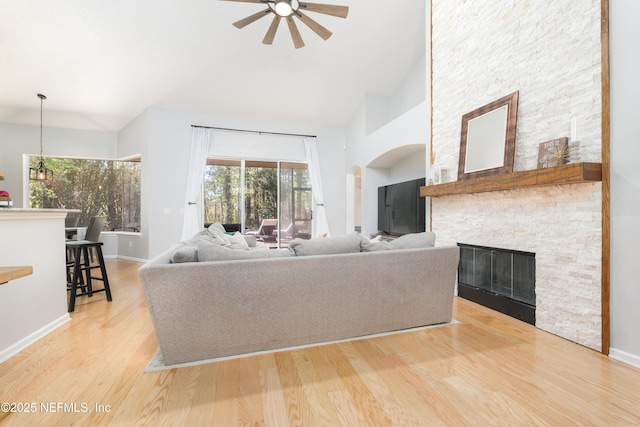 living room with light wood-type flooring, high vaulted ceiling, a ceiling fan, a stone fireplace, and baseboards