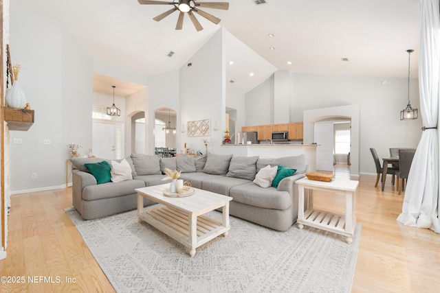 living room with high vaulted ceiling, ceiling fan with notable chandelier, arched walkways, light wood finished floors, and baseboards