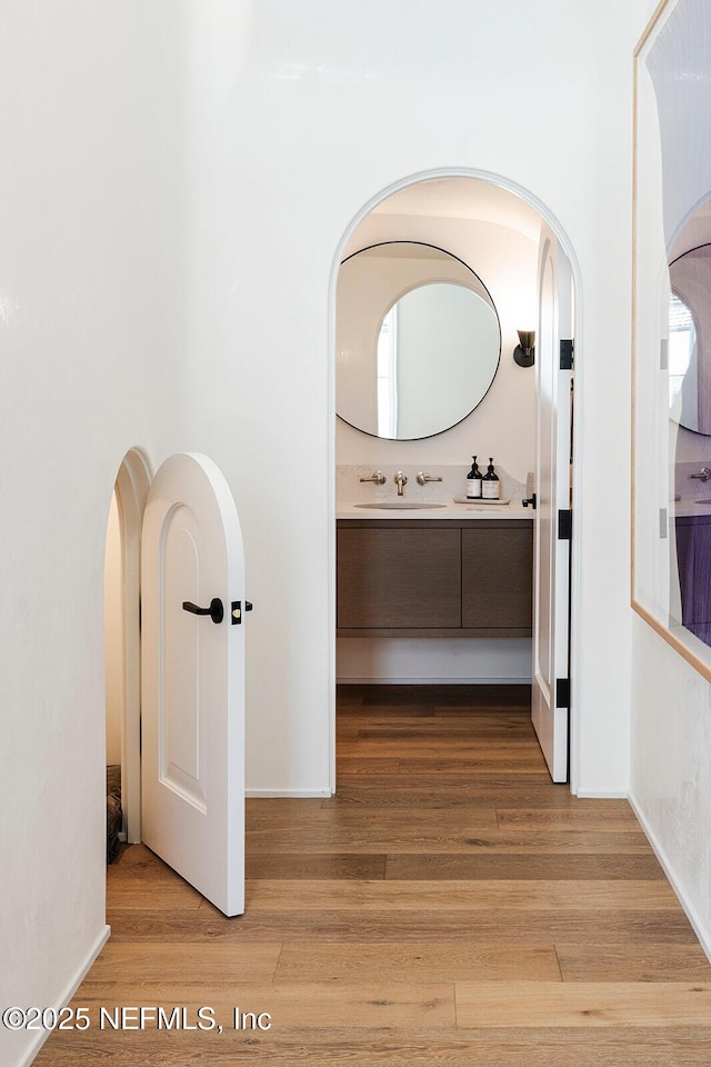 corridor featuring light wood-style floors, arched walkways, a sink, and baseboards