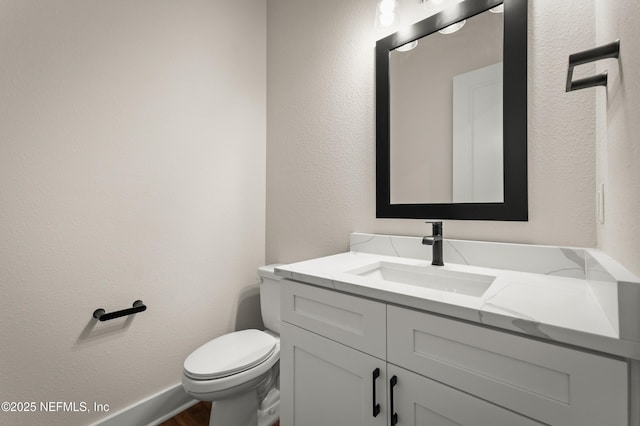 half bath featuring toilet, a textured wall, baseboards, and vanity