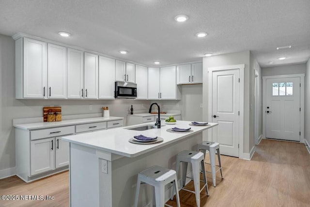 kitchen featuring stainless steel microwave, a sink, light wood-style flooring, and an island with sink