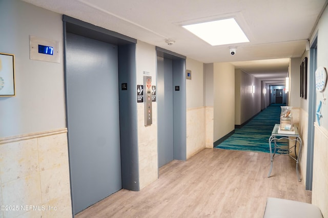 hallway featuring elevator, tile walls, wood finished floors, and a wainscoted wall