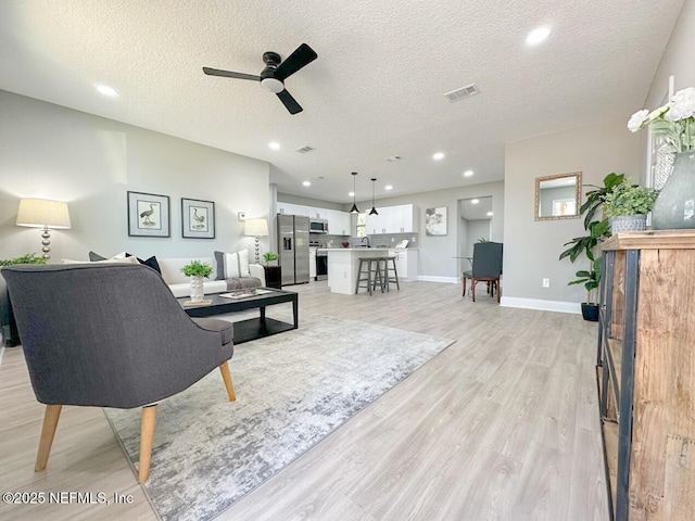 living area featuring baseboards, visible vents, a ceiling fan, a textured ceiling, and light wood-type flooring