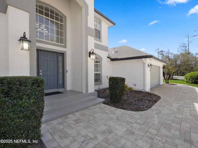 entrance to property with stucco siding, driveway, a garage, and roof with shingles