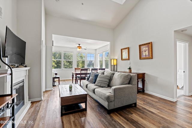 living room featuring a high ceiling, a glass covered fireplace, ceiling fan, wood finished floors, and baseboards