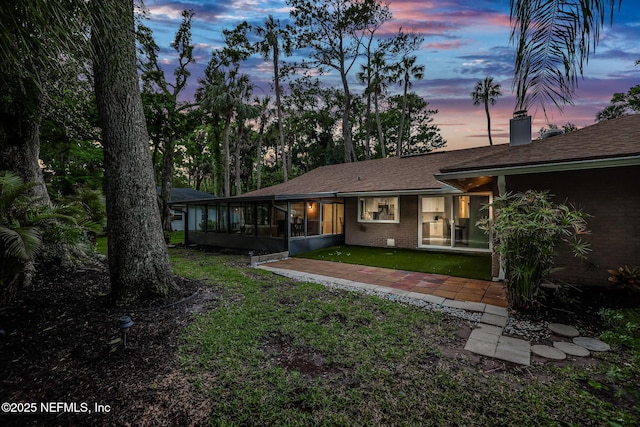 back of property at dusk with a sunroom, brick siding, a yard, and roof with shingles