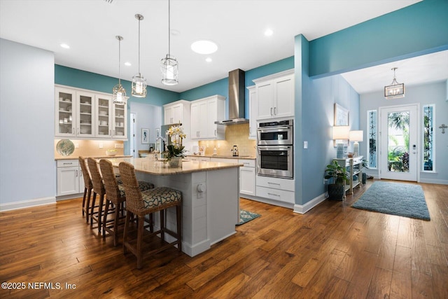 kitchen with a kitchen breakfast bar, dark wood-style flooring, stainless steel double oven, wall chimney range hood, and backsplash