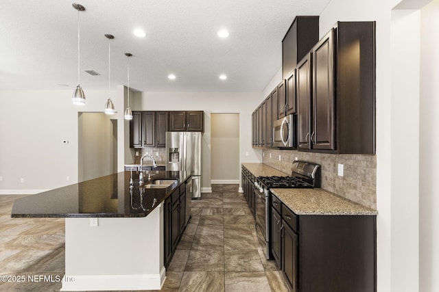 kitchen with an island with sink, a sink, dark stone countertops, stainless steel appliances, and dark brown cabinets