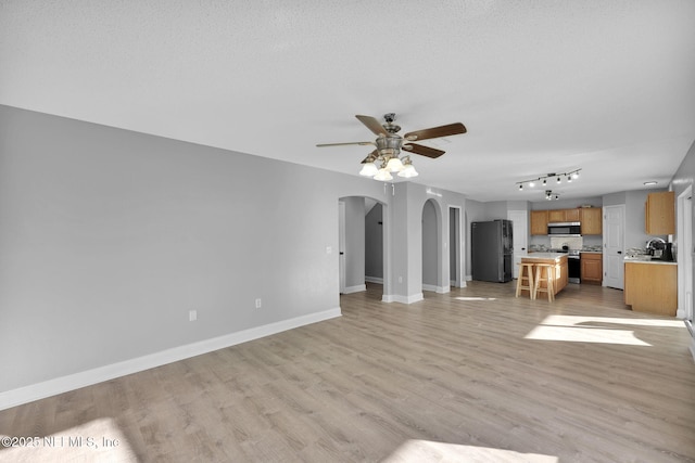 unfurnished living room with a textured ceiling, arched walkways, light wood finished floors, baseboards, and ceiling fan