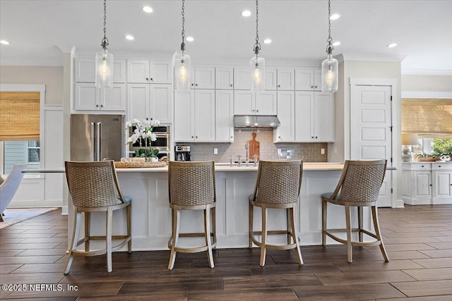 kitchen featuring dark wood-type flooring, under cabinet range hood, stainless steel appliances, white cabinetry, and a kitchen island with sink