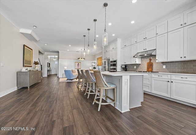 kitchen featuring backsplash, under cabinet range hood, ornamental molding, white cabinetry, and dark wood-style flooring