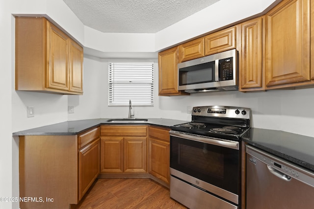 kitchen with a textured ceiling, stainless steel appliances, a sink, light wood-type flooring, and dark countertops