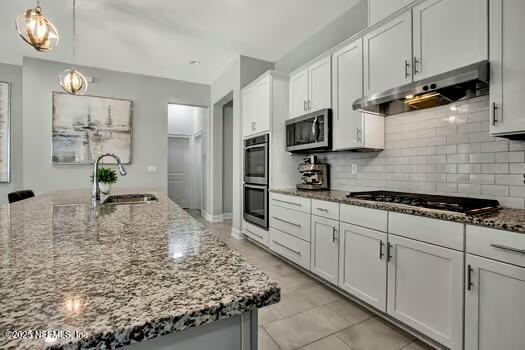 kitchen featuring dark stone counters, decorative backsplash, range hood, stainless steel appliances, and a sink