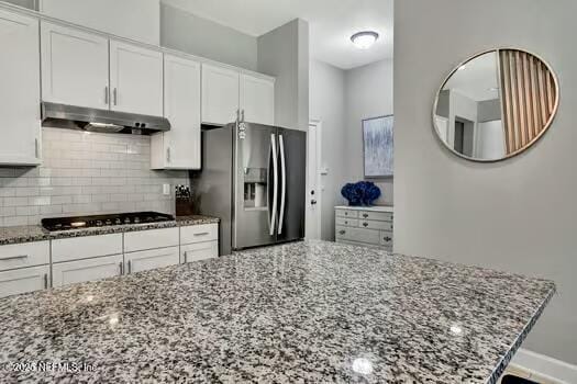 kitchen featuring black cooktop, dark stone counters, stainless steel refrigerator with ice dispenser, under cabinet range hood, and backsplash