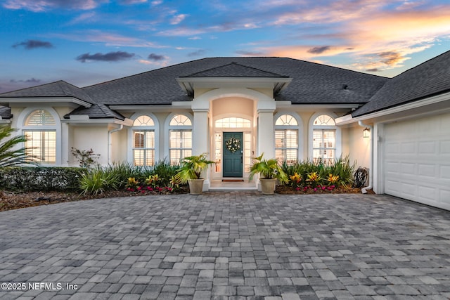 french country home featuring stucco siding, decorative driveway, roof with shingles, and an attached garage