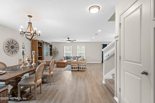 dining space featuring baseboards, light wood finished floors, stairs, a barn door, and ceiling fan with notable chandelier