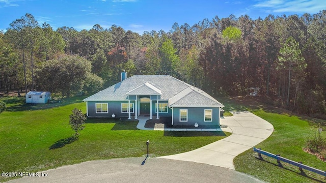 view of front of property with a front yard, a shingled roof, concrete driveway, an outdoor structure, and a view of trees