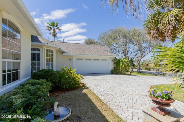 view of side of property featuring stucco siding, an attached garage, decorative driveway, and roof with shingles