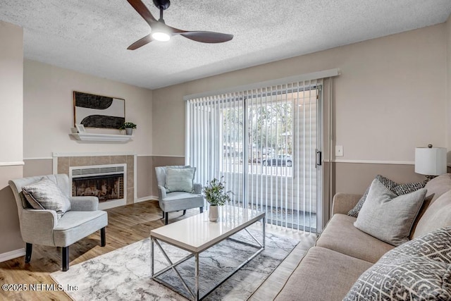 living room featuring a ceiling fan, a tiled fireplace, a textured ceiling, wood finished floors, and baseboards