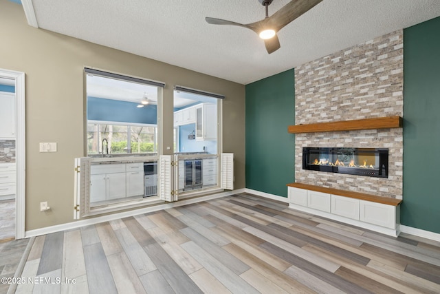 unfurnished living room with ceiling fan, wine cooler, light wood-style floors, a textured ceiling, and a large fireplace