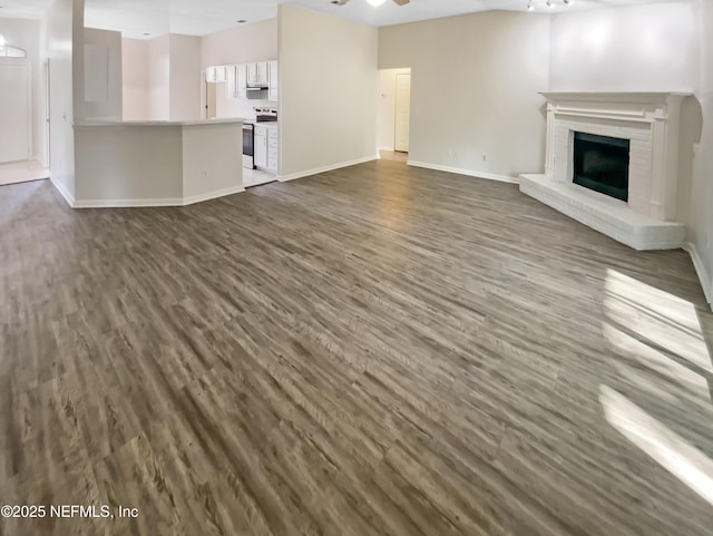 unfurnished living room with a brick fireplace, dark wood-style floors, baseboards, and ceiling fan