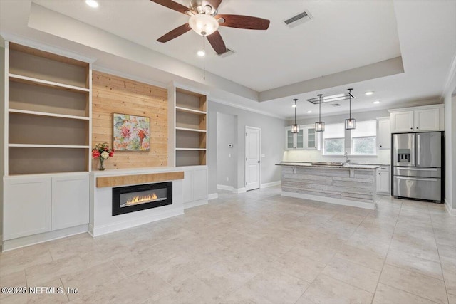 unfurnished living room featuring visible vents, built in features, a tray ceiling, a glass covered fireplace, and baseboards