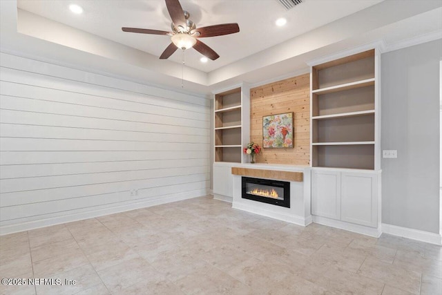 unfurnished living room featuring built in shelves, baseboards, a tray ceiling, ceiling fan, and a glass covered fireplace