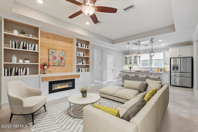 living area featuring built in features, baseboards, visible vents, a tray ceiling, and a glass covered fireplace