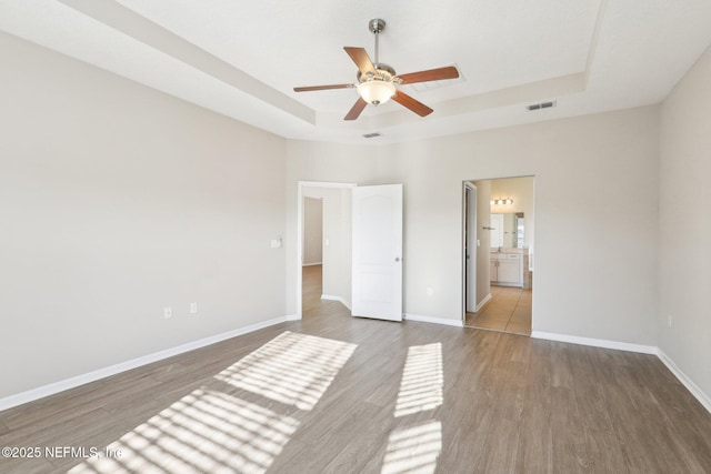 unfurnished bedroom featuring wood finished floors, visible vents, baseboards, a tray ceiling, and ceiling fan