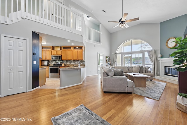 living room featuring baseboards, light wood-type flooring, a tile fireplace, high vaulted ceiling, and a ceiling fan