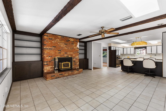 unfurnished living room featuring visible vents, beam ceiling, a wood stove, light tile patterned flooring, and a ceiling fan