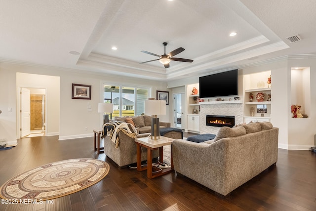 living area with visible vents, a tray ceiling, a glass covered fireplace, dark wood finished floors, and ceiling fan