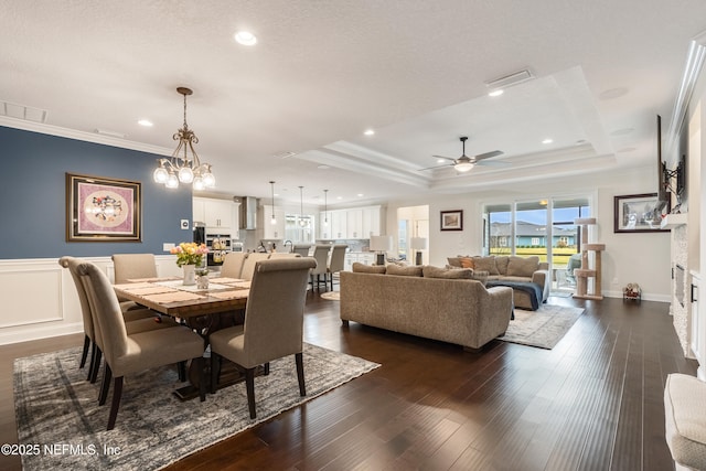 dining area with dark wood-type flooring, a wainscoted wall, ornamental molding, ceiling fan with notable chandelier, and a raised ceiling