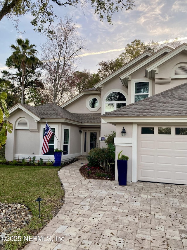 view of front facade with stucco siding, a shingled roof, decorative driveway, and a front lawn