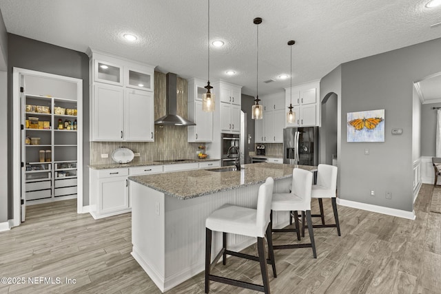 kitchen featuring wall chimney range hood, refrigerator with ice dispenser, light wood-style floors, arched walkways, and a sink