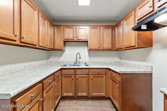 kitchen featuring under cabinet range hood, a sink, wood finished floors, brown cabinetry, and light countertops