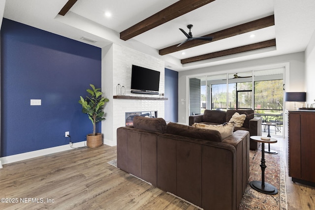 living room with light wood-type flooring, beamed ceiling, a large fireplace, baseboards, and ceiling fan