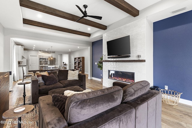 living area with light wood finished floors, visible vents, a large fireplace, beamed ceiling, and a ceiling fan