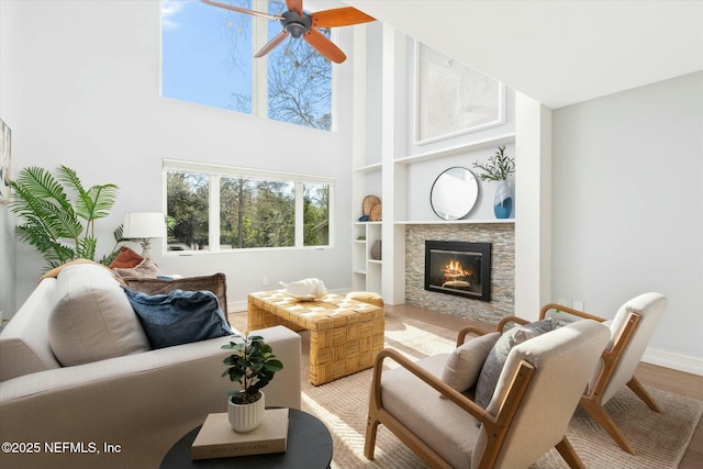 living area featuring wood finished floors, a towering ceiling, a stone fireplace, baseboards, and ceiling fan