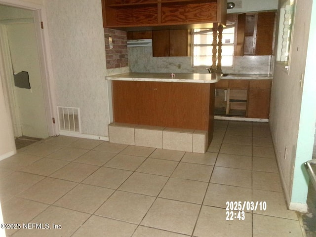 kitchen featuring visible vents, under cabinet range hood, tasteful backsplash, light tile patterned flooring, and brown cabinetry