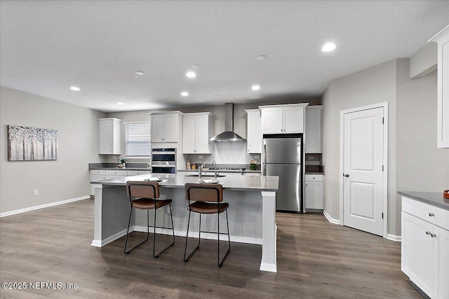 kitchen with stainless steel appliances, dark wood-style floors, a kitchen island with sink, and wall chimney range hood