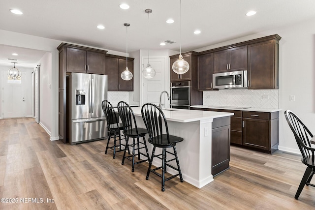 kitchen with light wood finished floors, dark brown cabinets, an island with sink, and appliances with stainless steel finishes